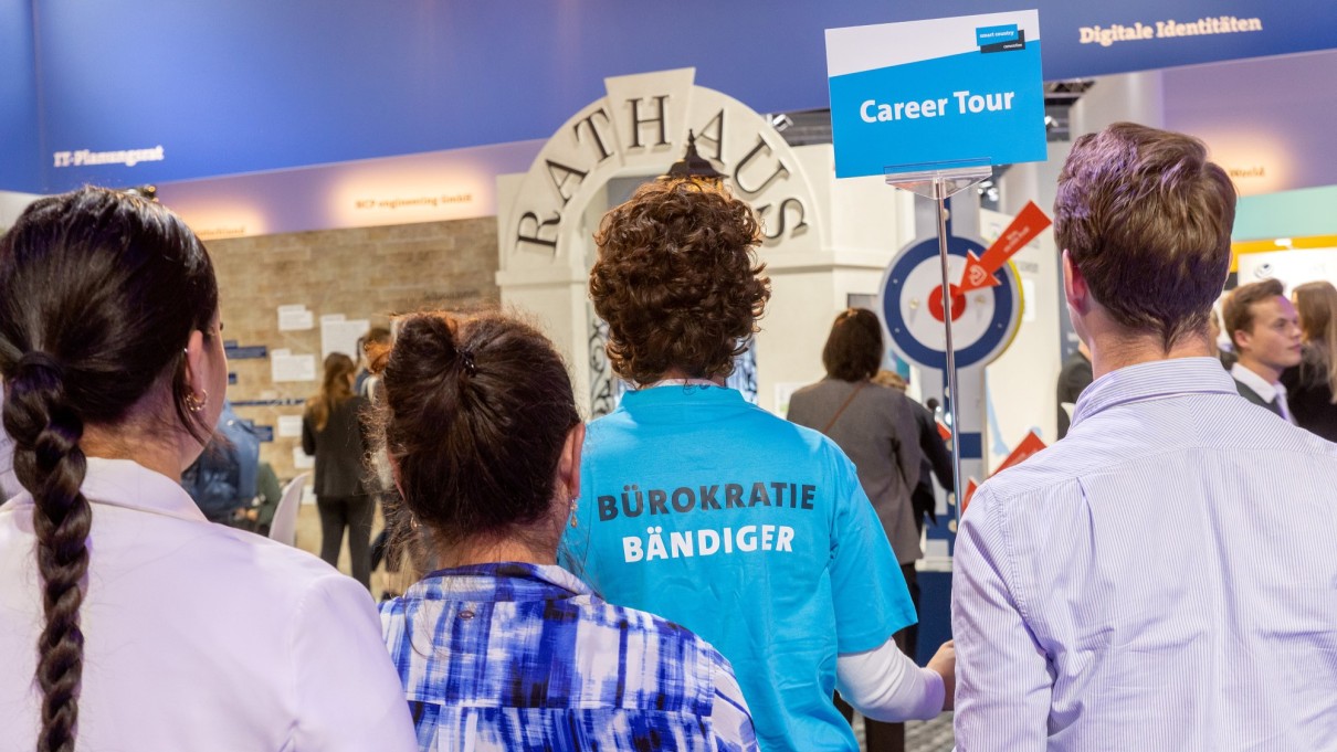 Vier Personen von hinten auf dem Messestand des Bundesministeriums des Innern und für Heimat, eine Person hält ein Schild mit der Aufschrift „Career Tour“ Vier Personen von hinten auf dem Messestand des Bundesministeriums des Innern und für Heimat, eine Person hält ein Schild mit der Aufschrift „Career Tour“