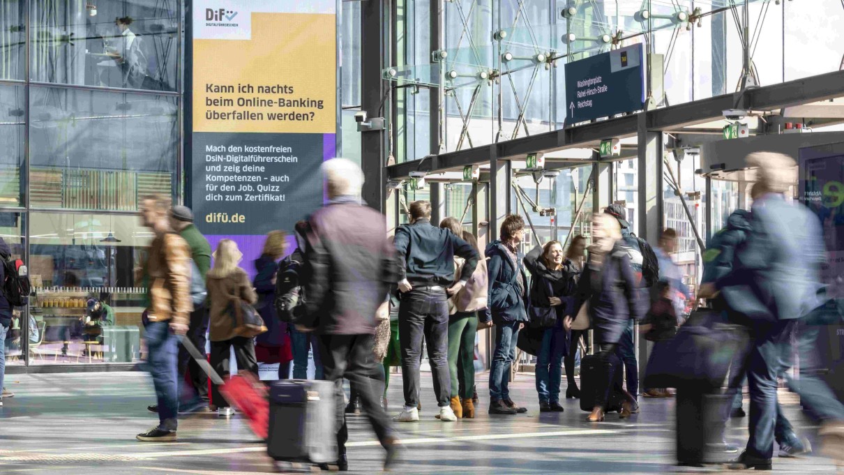 Menschen am Bahnhof, im Hintergrund eine großflächige Werbung zum DiFü