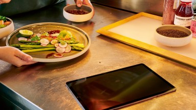 Links ein Teller mit Gemüse, rechts vorn ein Tablet, im Hintergrund Soßen und Gewürze 