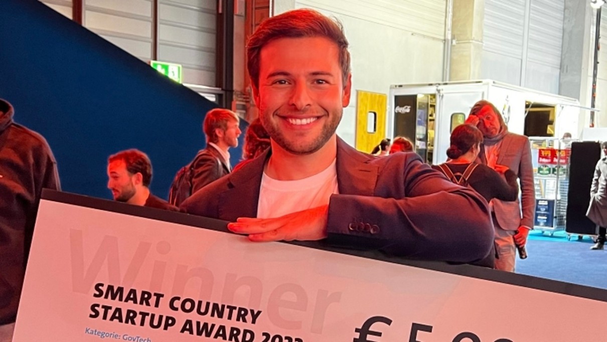 Junger Mann im Vordergrund, der einen großen Check mit dem Preisgeld für den Smart Country Startup Award in der Hand hält. 