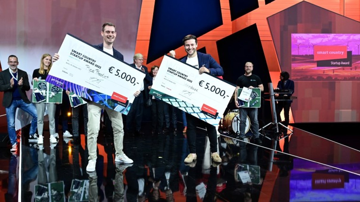 Auf der Bühne stehen zwei Männer stolz und präsentieren die großen Schecks mit den 5.000 Euro Preisgeld in ihren Händen.