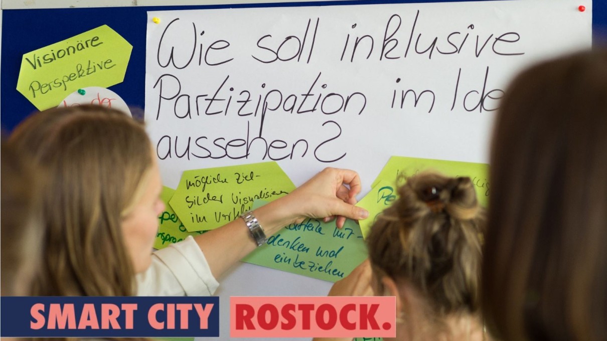 Drei Frauen vor einer Flipchart, auf der die Frage „Wie soll inklusive Partizipation aussehen?“ steht. Im Vordergrund der Slogan „Smart City Rostock. Deine Stadt von Morgen.“ 