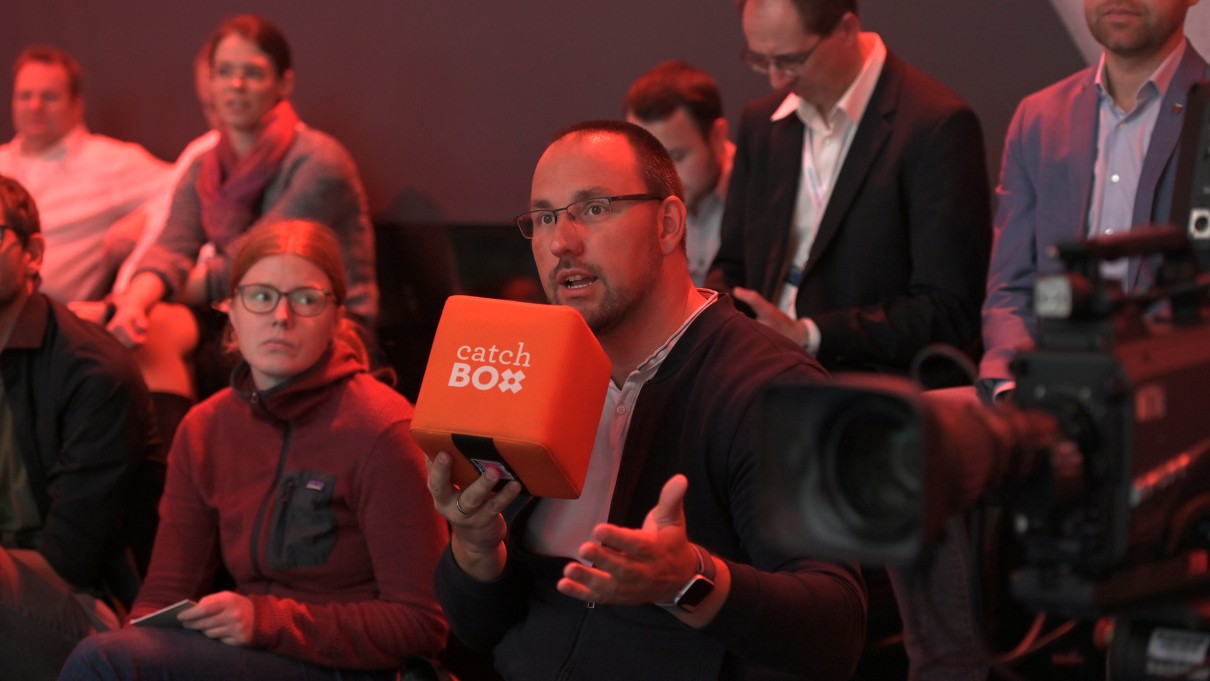 Eine Gruppe von Zuschauerinnen und Zuschauern ist abgebildet. Ein Mann hält ein Mikrofon in der Hand.