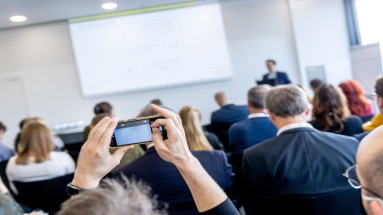 Workshop, speaker is in the background with a presentation on the wall, the audience is in the foreground. 