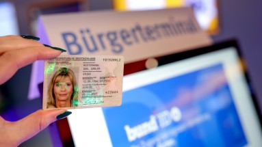 In the foreground, a hand holding an ID card. In the background a citizen terminal for the BundID.