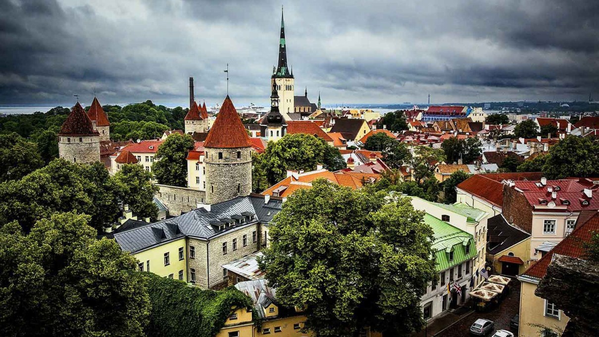 View of the Old Town of Tallinn, Estonia, with medieval towers, red roofs and green trees standing out under a dramatically cloudy sky.
