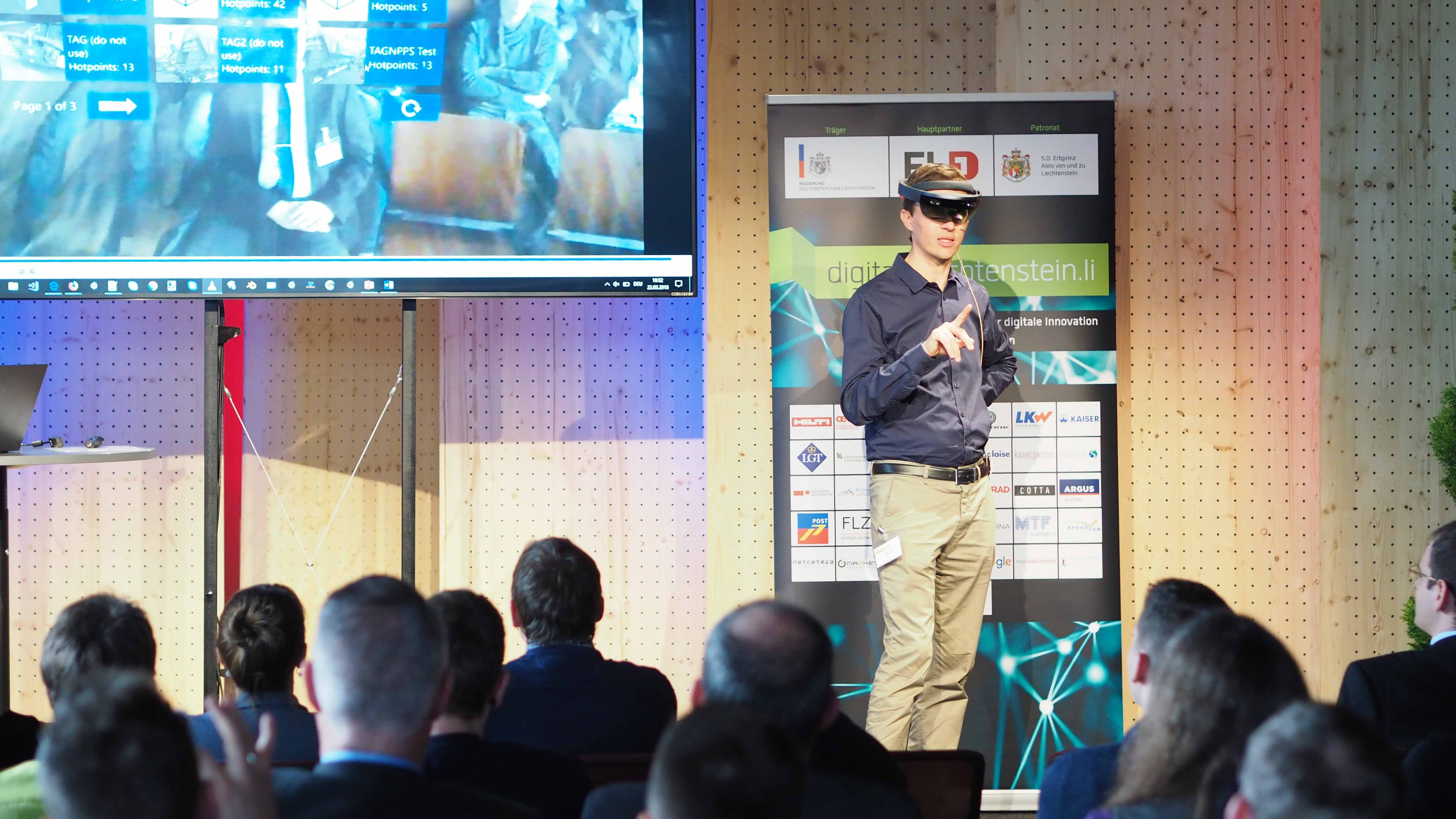 A man presents virtual reality glasses, in the foreground the audience can be seen from behind, on the wall a projection.