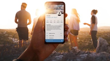 Das Bild zeigt eine Gruppe von Menschen vor einem Sonnenuntergang, eine Hand hält ein Smartphone mit offener Community-App.
