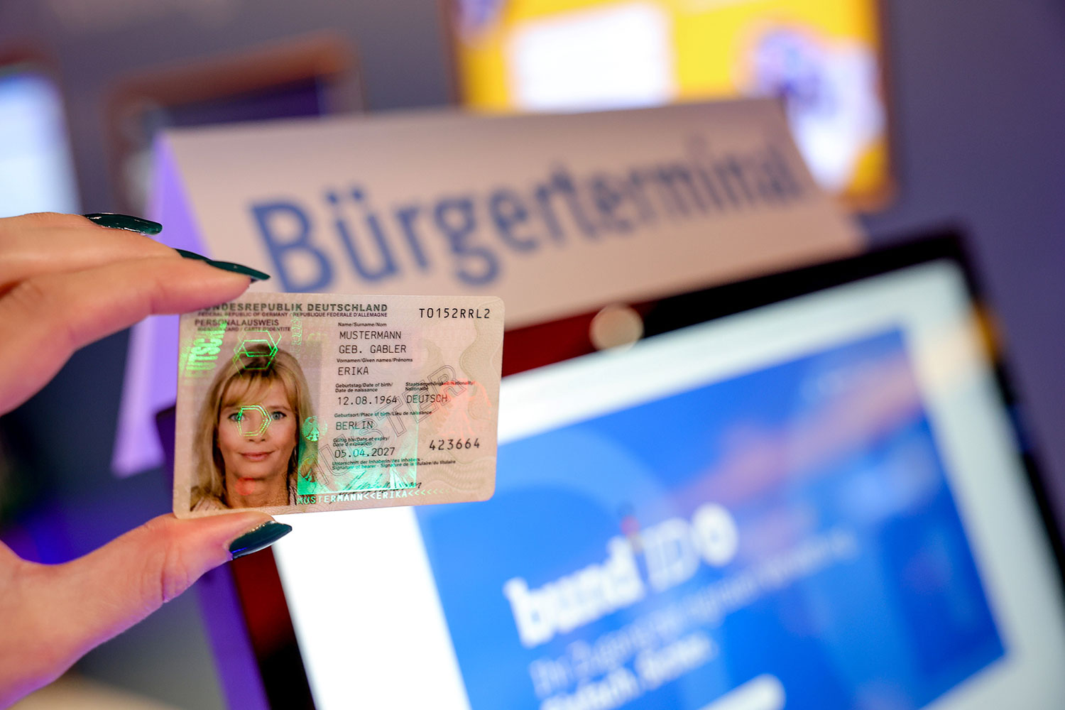 Im Vordergrund eine Hand, die einen Personalausweis hält. Im Hintergrund ein Bürgerterminal zur BundID.