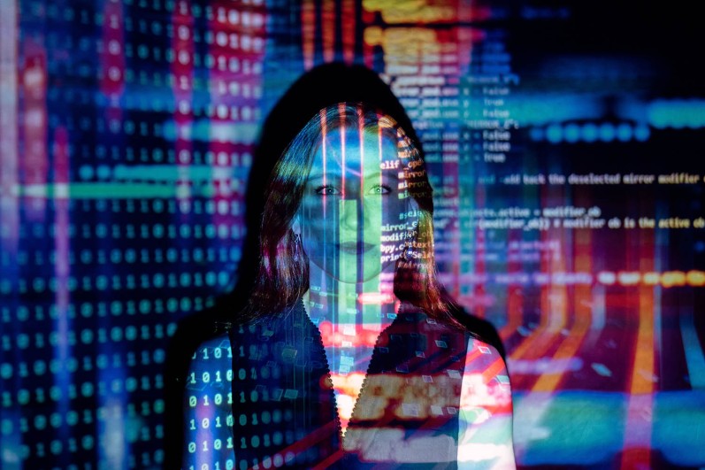 Frau mit projiziertem farbigem Code im Hintergrund. Symbolbild für Informationssicherheit und Datenschutzrisiken in der Smart City.